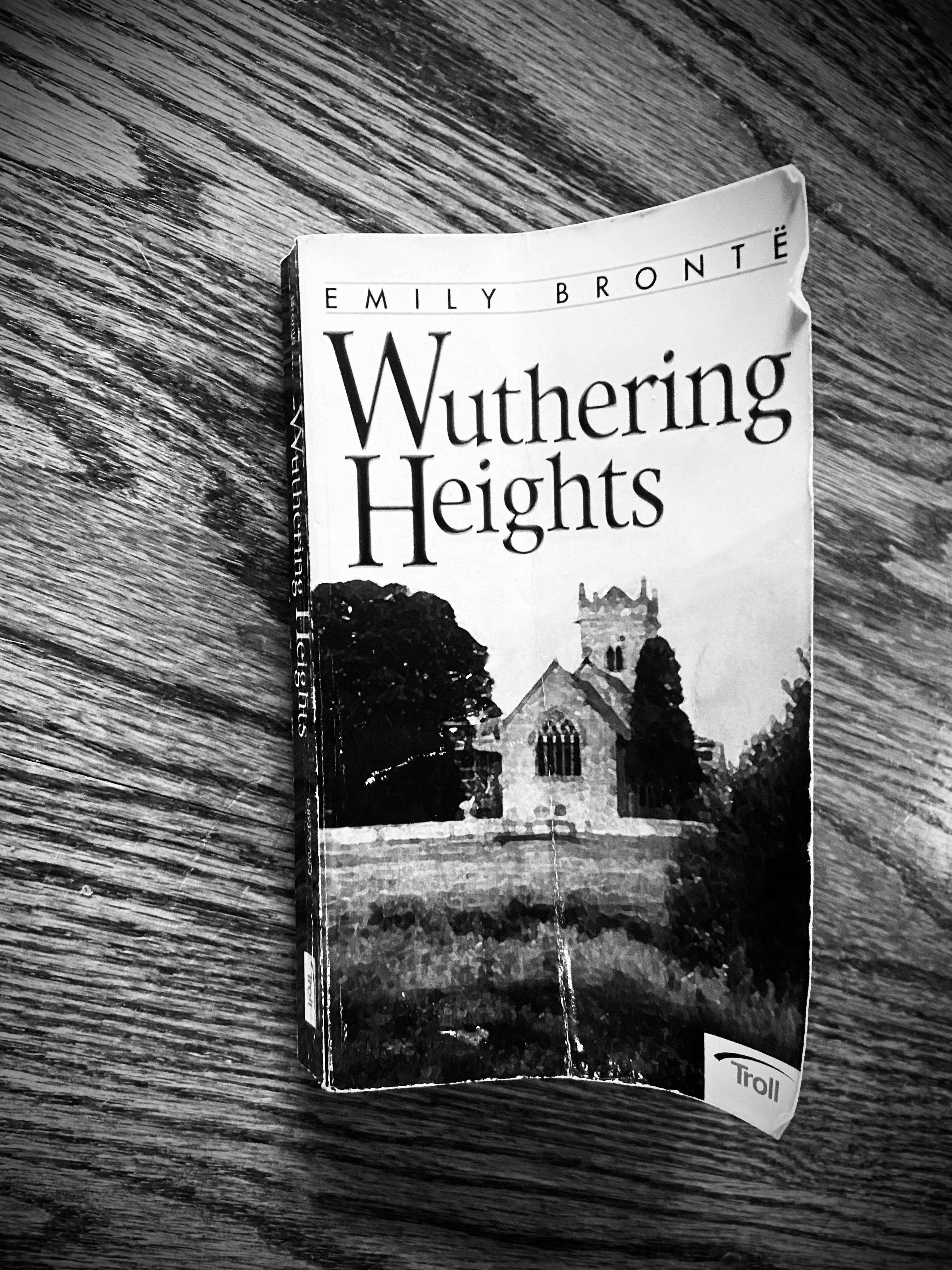 black and white photograph of a wooden tabletop with a mass market paperback copy of Emily Brontë's Wuthering Heights face up on it.